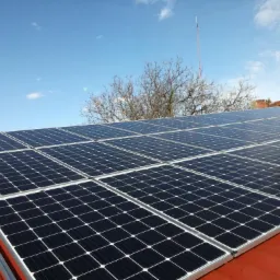 Rząd paneli słonecznych na czerwonym dachu budynku w słoneczny dzień, widoczne drzewo bez liści i antena w tle.