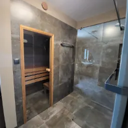 Nowoczesna łazienka z kabiną prysznicową typu walk-in i sauną z drewnianymi ławkami, wykończona szarymi płytkami wielkoformatowymi.