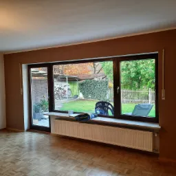 Pomieszczenie po malowaniu ścian na brązowo, widoczny parkiet, grzejnik pod oknem i widok na ogród z meblami ogrodowymi.