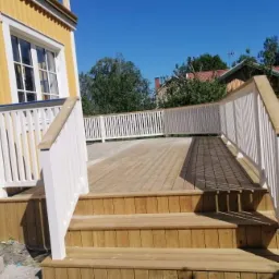 Drewniany taras z białymi balustradami przy żółtym domu z widocznymi schodami i kostką brukową na ziemi.