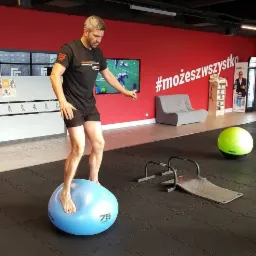 Mężczyzna w czarnej koszulce i spodenkach ćwiczy równowagę, stojąc boso na niebieskiej piłce Bosu w siłowni z czerwoną ścianą i napisem #możeszwszystko.