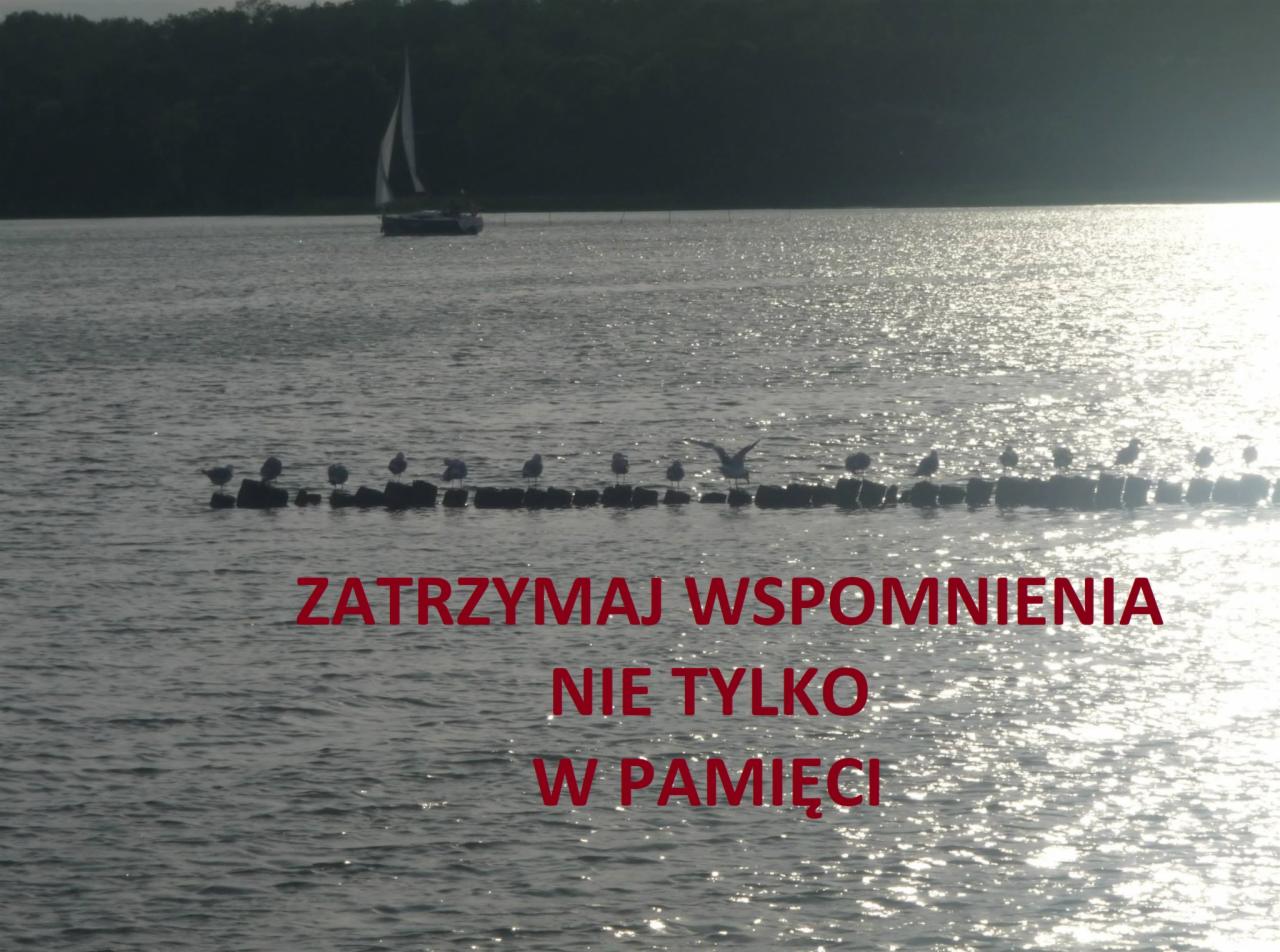 Krajobraz morski z żaglówką w oddali, rzędem ptaków siedzących na pirsie i napisem 'Zatrzymaj wspomnienia nie tylko w pamięci' nałożonym na zdjęcie.