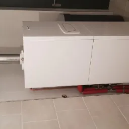 Nowoczesny, biały piec gazowy z chromowanym kominem odprowadzającym spaliny, zamontowany w pomieszczeniu wyłożonym jasnymi, kwadratowymi płytkami. Widoczne czerwone rury instalacji grzewczej...