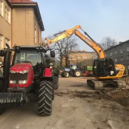 Czerwony traktor Massey Ferguson, żółta koparka JCB i niebieski traktor New Holland w tle, podczas prac ziemnych przy budynku.