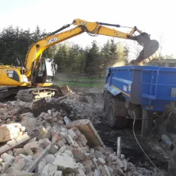 Żółta koparka JCB załadowuje gruz do niebieskiej ciężarówki na terenie rozbiórki, w tle widoczne drzewa.