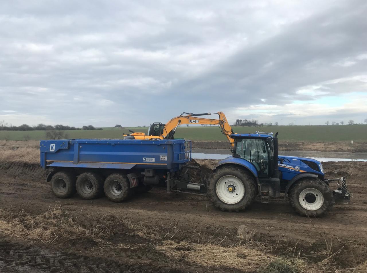 Żółta koparka JCB ładuje ziemię do niebieskiej przyczepy ciągniętej przez niebieski ciągnik New Holland na polu z widocznym zbiornikiem wodnym w tle.