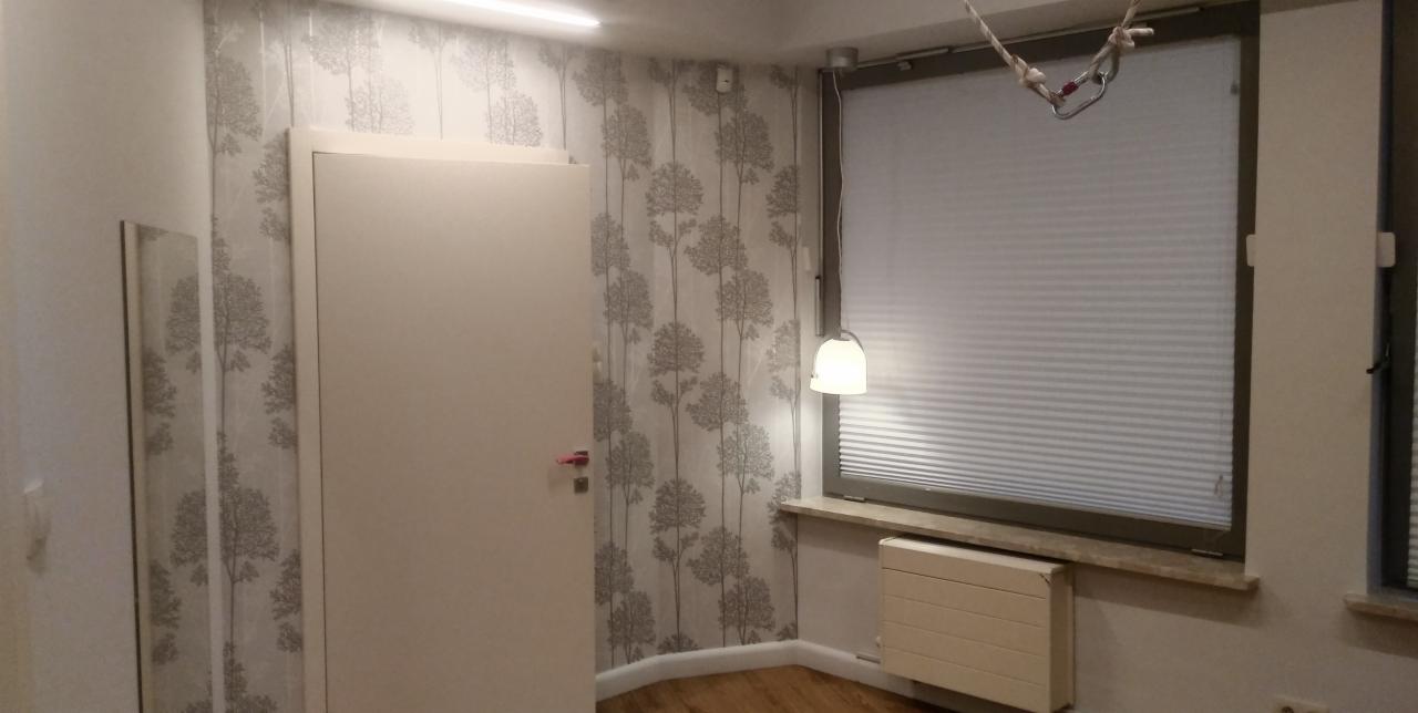 Pokój po remoncie z tapetą w drzewa, białe drzwi, okno z żaluzjami plisowanymi, grzejnik i lampa wisząca.