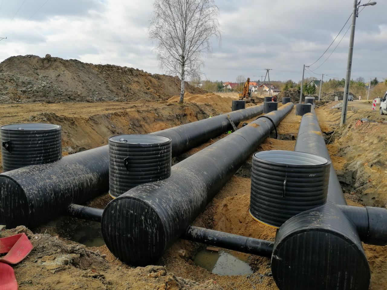 Instalacja podziemnej infrastruktury kanalizacyjnej z rur karbowanych i studzienek rewizyjnych na placu budowy, widoczny wykop z ziemią, w tle koparka.