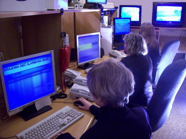 Stanowisko dyspozytorskie z kilkoma osobami monitorującymi ekrany z napisem 'Krajowa Sieć Alarmowa SKSR', telefony na biurkach, monitory CCTV w tle.