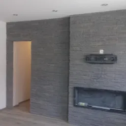 Ściana w salonie wykończona szarą okładziną kamienną, z przygotowanym miejscem na kominek i instalację TV. Widoczny fragment podłogi z paneli oraz wejście do innego pomieszczenia.