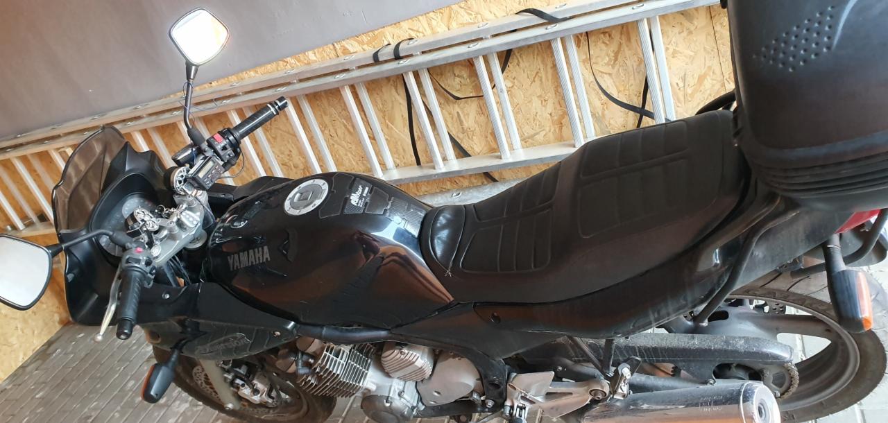 Czarny motocykl Yamaha w garażu, widoczna drabina oparta o ścianę, detale motocykla: kierownica, siedzenie, silnik.