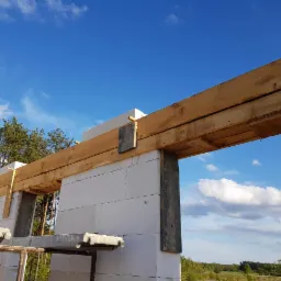 Wznoszenie ścian z bloczków białych na budowie domu w Białymstoku, widoczne drewniane nadproża okienne i rusztowanie, błękitne niebo w tle.