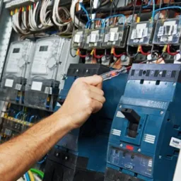 Elektryk sprawdza napięcie w rozdzielnicy za pomocą próbnika napięcia; widoczne liczniki energii elektrycznej, przewody i bezpieczniki.