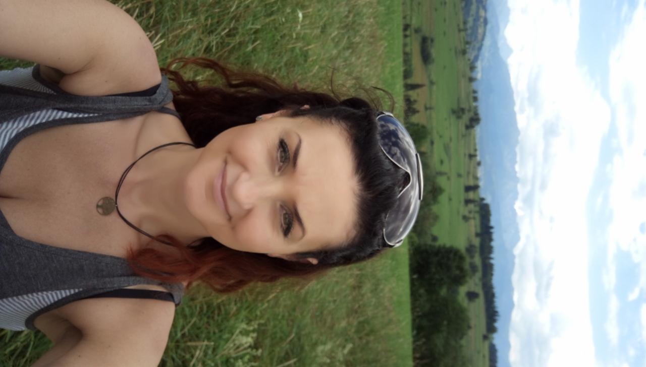 Uśmiechnięta kobieta z okularami na głowie robi selfie na tle zielonych wzgórz i błękitnego nieba z chmurami.