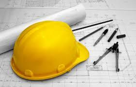 Żółty kask budowlany, zwinięty plan architektoniczny, linijka, ołówek, pisaki i cyrkiel na tle rysunku technicznego.