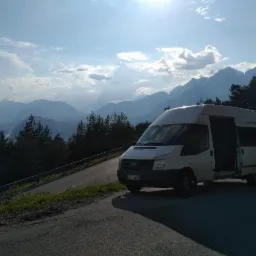 Biały bus Ford Transit z otwartymi drzwiami bocznymi zaparkowany na poboczu drogi z widokiem na dolinę i góry w tle, słoneczny dzień.