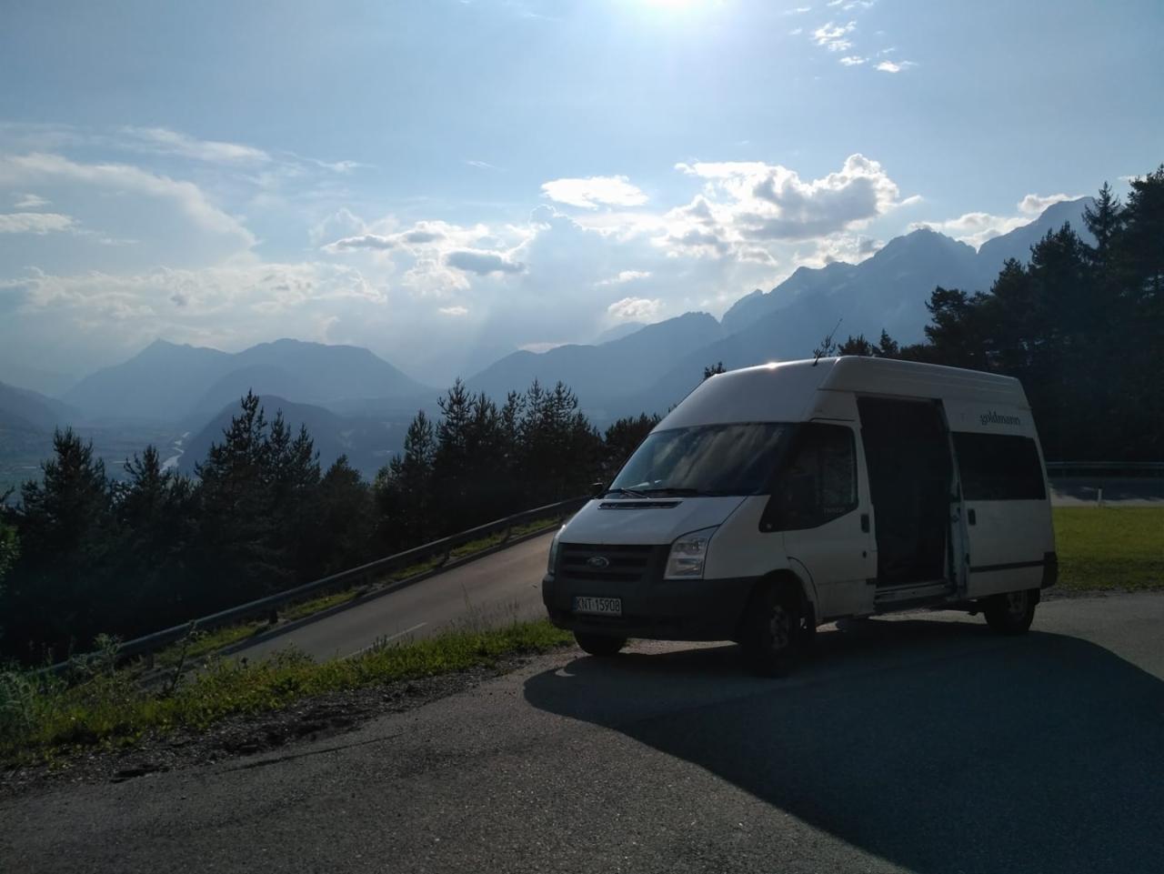 Biały bus Ford Transit z otwartymi drzwiami bocznymi zaparkowany na poboczu drogi z widokiem na dolinę i góry w tle, słoneczny dzień.