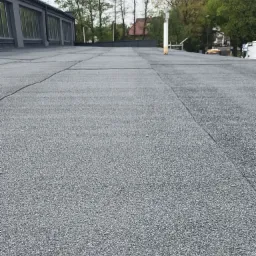 Szczelny dach pokryty papą termozgrzewalną w Warszawie, z widocznymi łączeniami i kominem wentylacyjnym. Widok z niskiej perspektywy, budynki w tle.