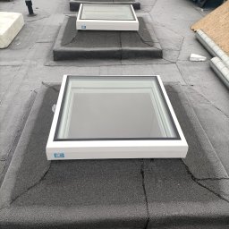 SABER INVEST Sp. zo.o - Montaż okien dachowych na pokryciu z papy. Widoczne trzy okna w białych ramach, układane w rzędzie w kierunku oddalającym się od obserwatora.