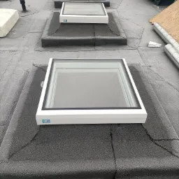 Montaż okien dachowych na pokryciu z papy. Widoczne trzy okna w białych ramach, układane w rzędzie w kierunku oddalającym się od obserwatora.