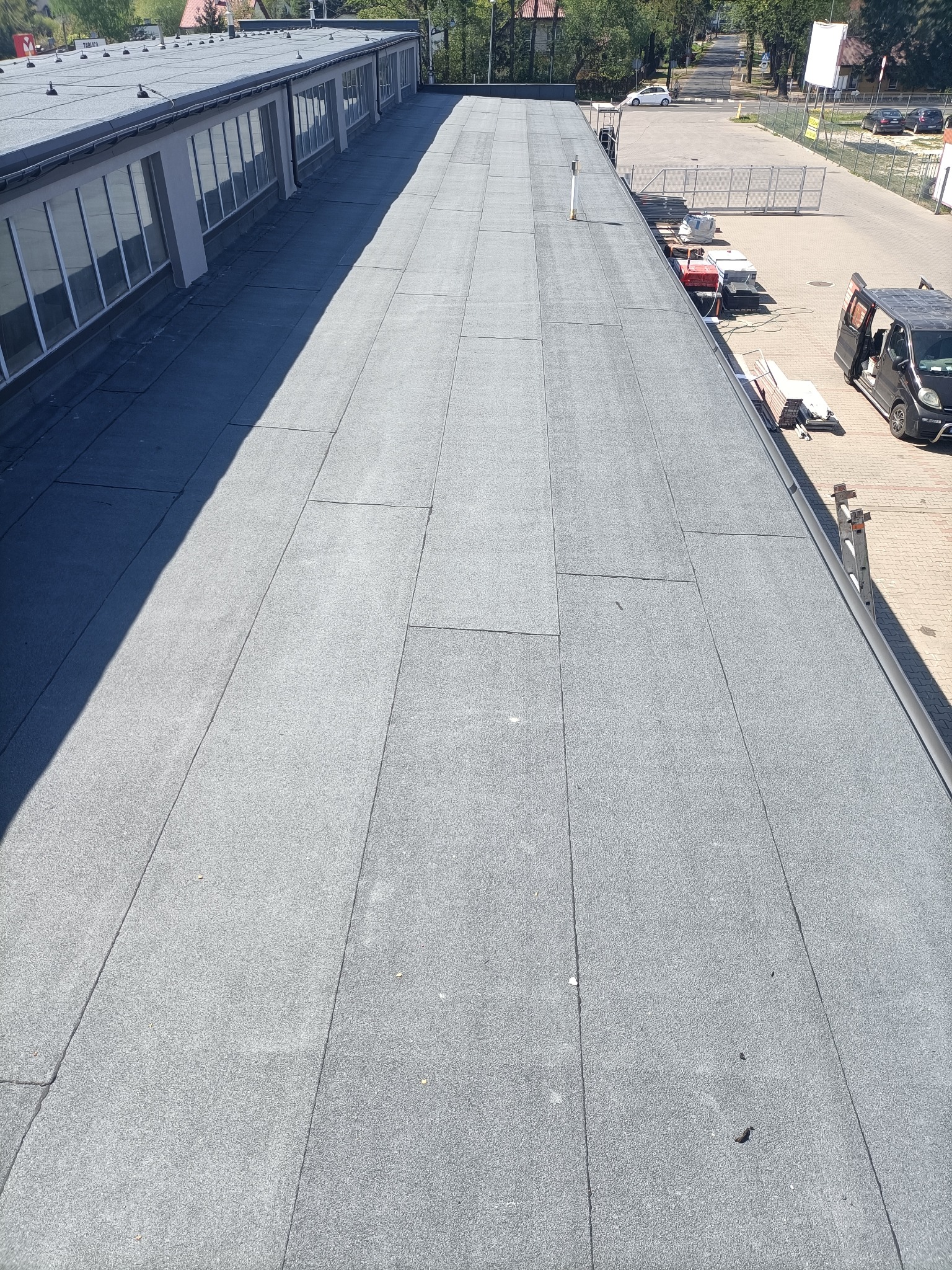 Szary, płaski dach pokryty papą termozgrzewalną na budynku przemysłowym, widok z góry. Na dachu widoczne łączenia papy. W tle zaparkowane auto i drzewa.