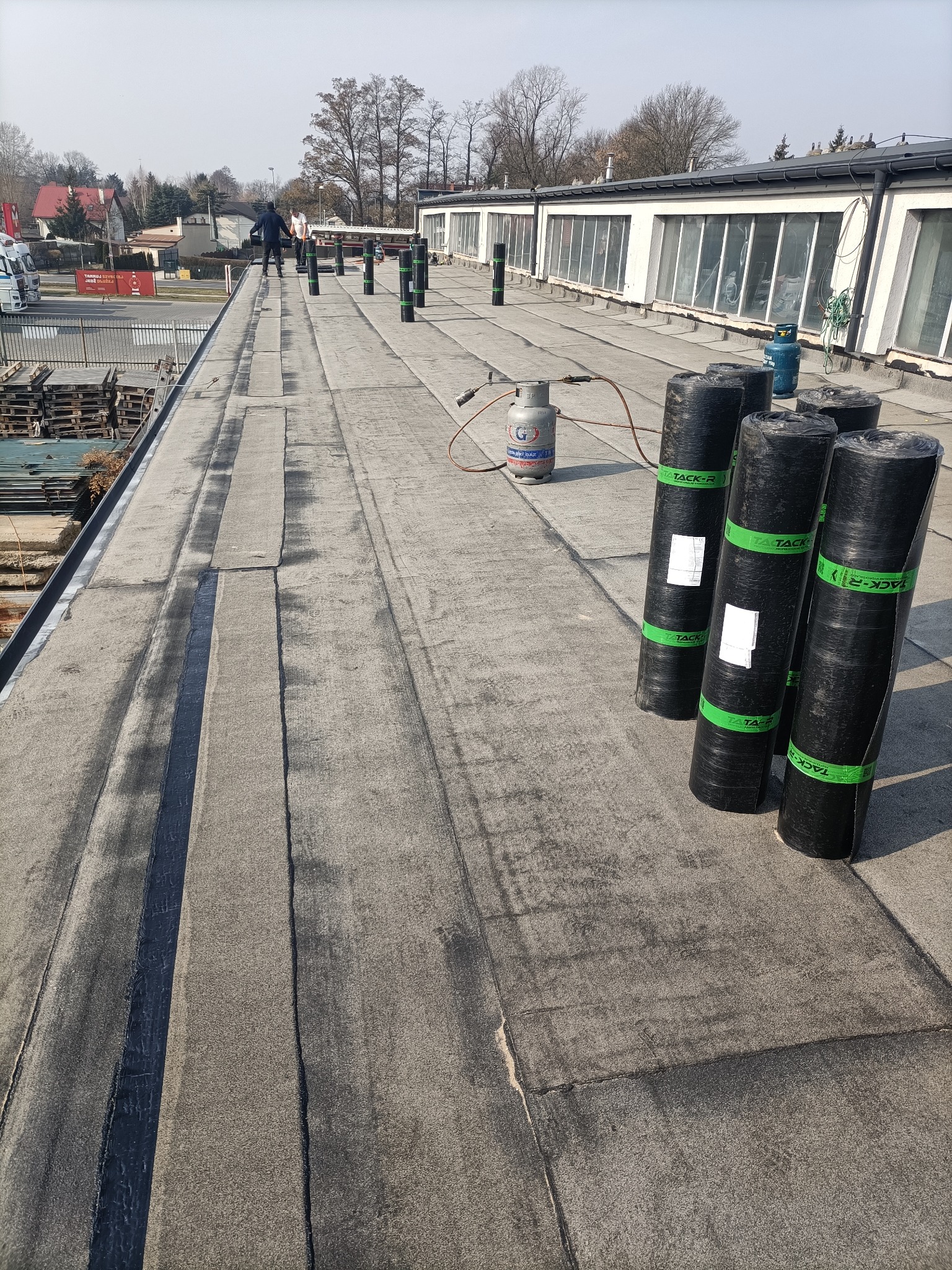 Prace dekarskie na płaskim dachu: zgrzewanie papy, widoczne rolki papy Tack-R, butle z gazem i dekarz w oddali. Dach pokryty papą w szarym kolorze.