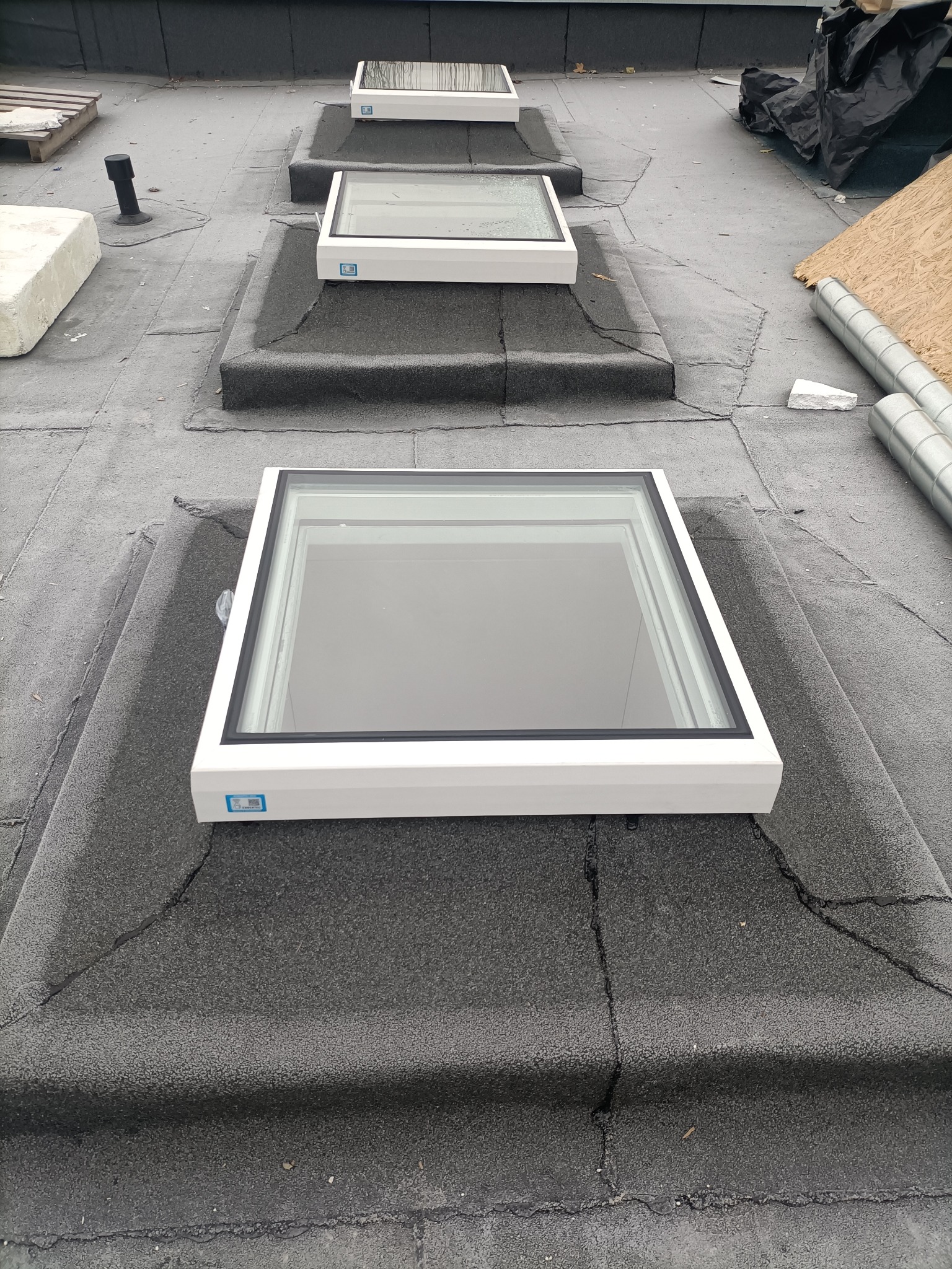 Montaż okien dachowych na pokryciu z papy. Widoczne trzy okna w białych ramach, układane w rzędzie w kierunku oddalającym się od obserwatora.