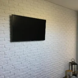 Ściana wykończona białymi płytkami imitującymi cegłę, z zawieszonym telewizorem LCD, stolik dekoracyjny z lampionem i elementami dekoracyjnymi w rogu pokoju.