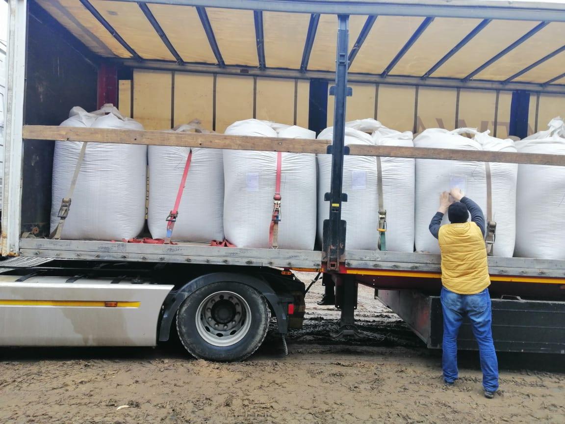 Mężczyzna w żółtej kamizelce zabezpiecza ładunek worków w naczepie ciężarówki, widoczne pasy mocujące i drewniane podpory.