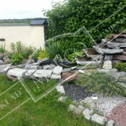 Ogród skalny z kaskadą wodną zbudowaną z nieregularnych, szarych płyt kamiennych, otoczony roślinnością i oświetlony lampami solarnymi; w tle mur i zielona ściana z krzewów.