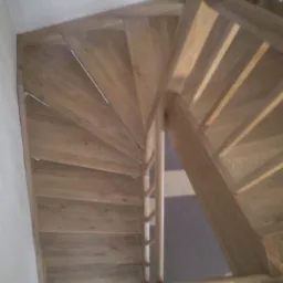 Widok z góry na kręte schody z jasnego drewna, z widoczną konstrukcją wsporczą i fragmentem ściany w tle.