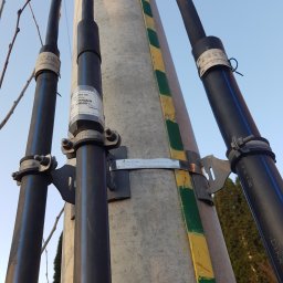 EMI GROUP - Słup energetyczny z przymocowanymi przewodami w czarnej osłonie, zabezpieczonymi obejmami. Widoczne metalowe mocowania i oznaczenia na przewodach.