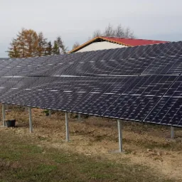 Instalacja fotowoltaiczna na metalowych podporach, widoczny traktor w tle. Panele skierowane w stronę słońca, drabina oparta o konstrukcję. Realizacja w wiejskim otoczeniu.
