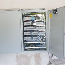 EMI GROUP - Otwarta skrzynka rozdzielcza z widocznymi zabezpieczeniami, przewodami i elementami montażowymi na białej ścianie. Na drzwiach wentylator i schemat.