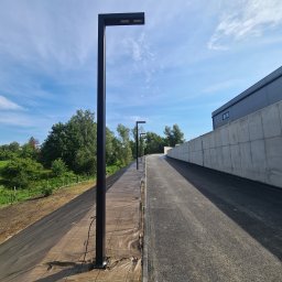 EMI GROUP - Szpaler nowoczesnych, czarnych lamp parkowych wzdłuż ścieżki, z widocznym okablowaniem. Tło: zieleń i budynek. Błękitne niebo z chmurami w kadrze.