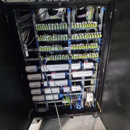 EMI GROUP - Otwarta szafa sterownicza z licznymi przewodami, listwami zaciskowymi, zabezpieczeniami i szynami montażowymi. Widoczne kable w różnych kolorach ułożone wewnątrz szafy.