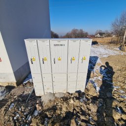 EMI GROUP - Biała, modułowa stacja transformatorowa Inoccbox 52959/RD-3, z żółtymi znakami ostrzegawczymi, usytuowana na placu budowy z koparką w tle.