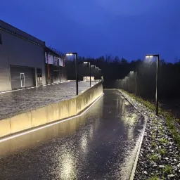 Oświetlona ścieżka wzdłuż budynku z lampami i podświetleniem LED, mokra nawierzchnia odbija światło, kamienny brzeg z roślinami, pochmurne niebo.