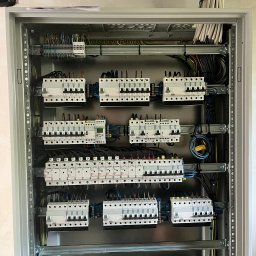 EMI GROUP - Rozdzielnica elektryczna w szarej obudowie, widoczne zabezpieczenia, przewody i szyny montażowe. Uporządkowany układ kabli na górze skrzynki.