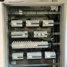 Rozdzielnica elektryczna w szarej obudowie, widoczne zabezpieczenia, przewody i szyny montażowe. Uporządkowany układ kabli na górze skrzynki.