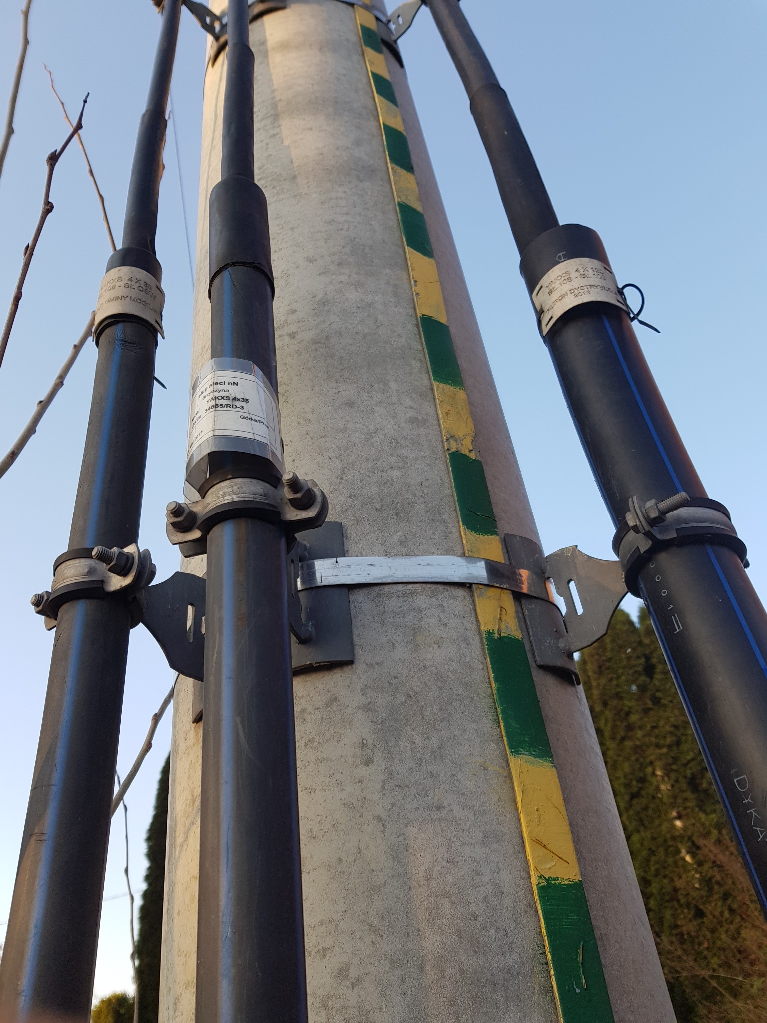 Słup energetyczny z przymocowanymi przewodami w czarnej osłonie, zabezpieczonymi obejmami. Widoczne metalowe mocowania i oznaczenia na przewodach.