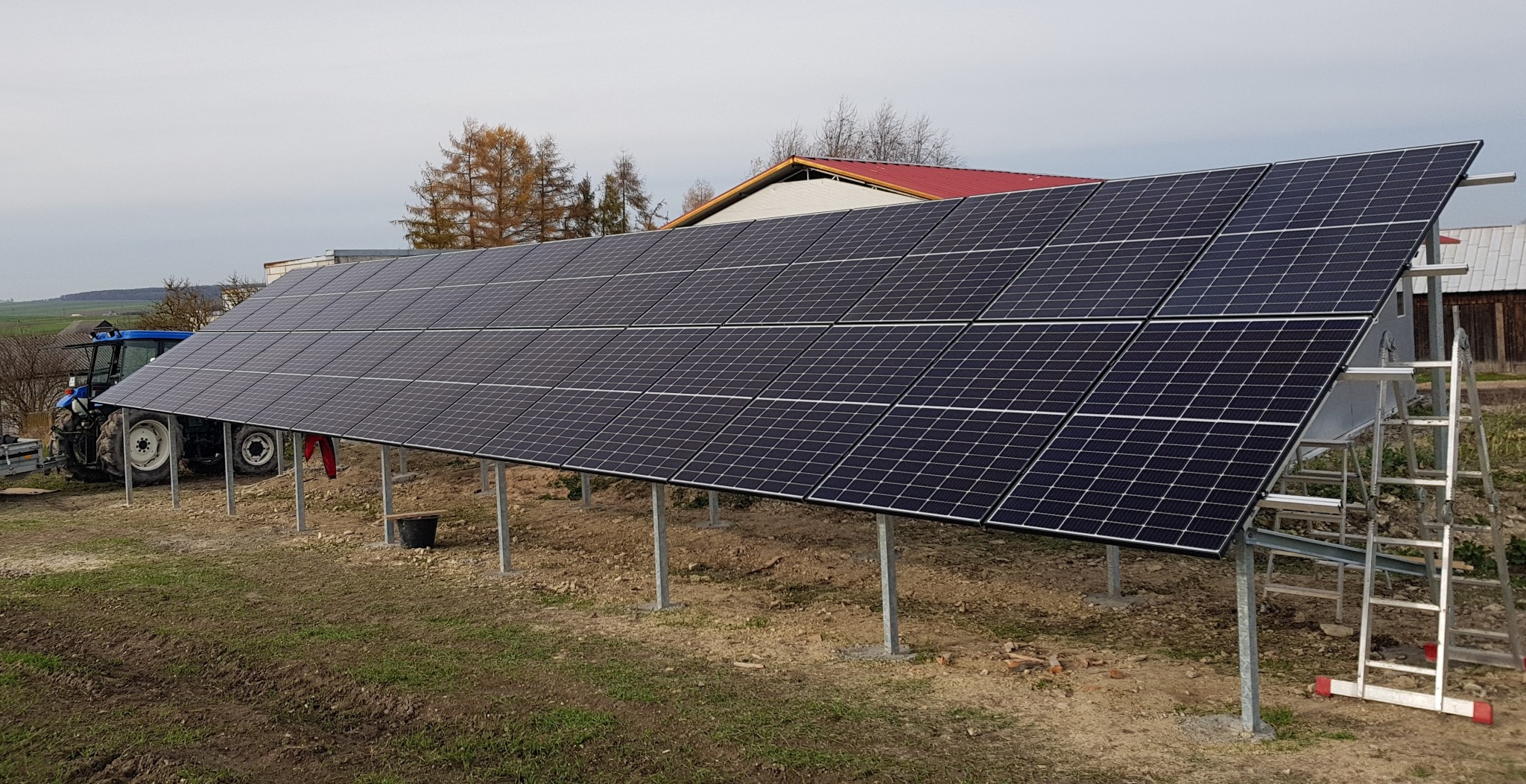 Instalacja fotowoltaiczna na metalowych podporach, widoczny traktor w tle. Panele skierowane w stronę słońca, drabina oparta o konstrukcję. Realizacja w wiejskim otoczeniu.