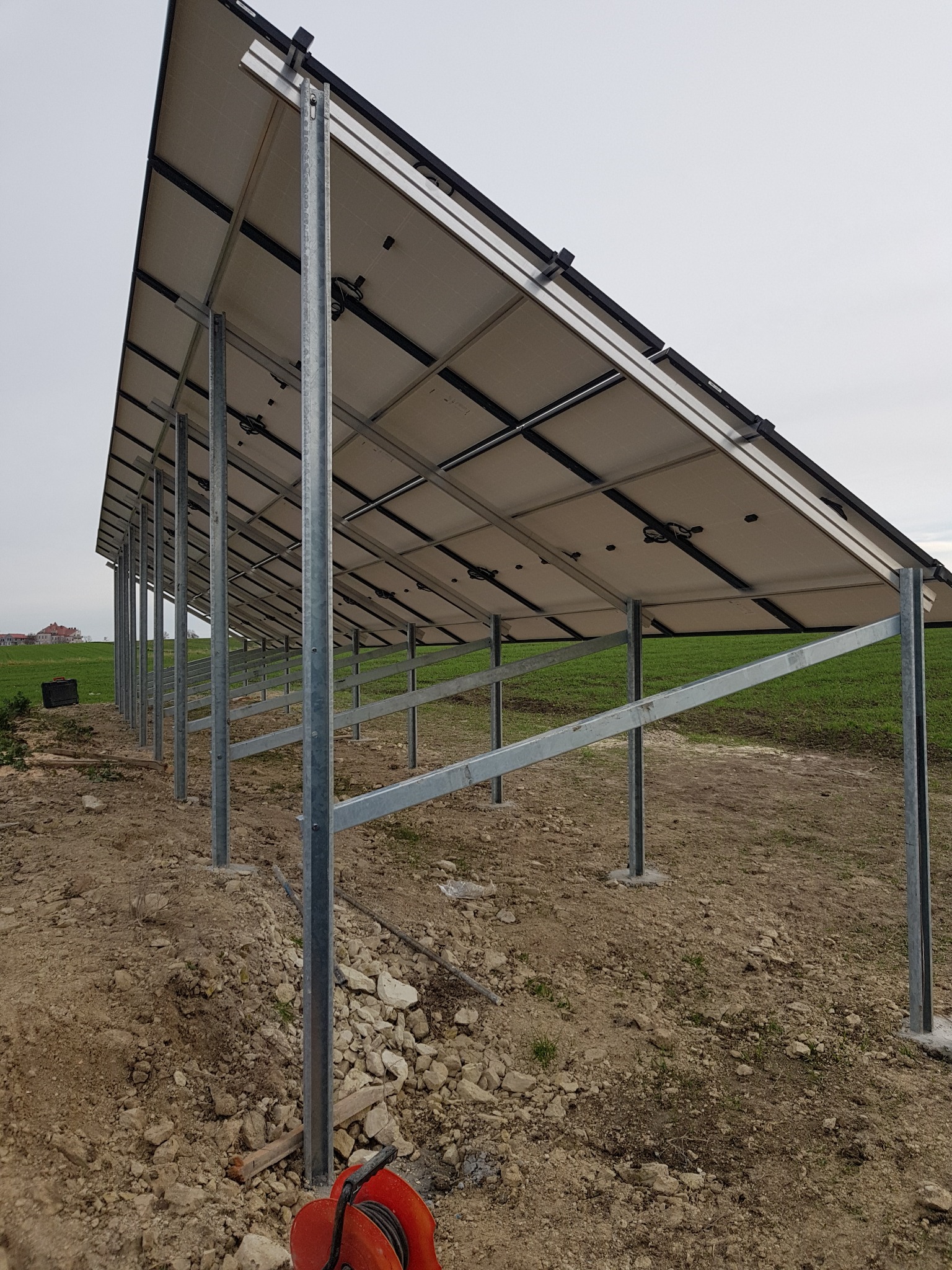 Montaż paneli fotowoltaicznych na metalowej konstrukcji, widok od spodu, z ziemią i zielenią w tle, kabel w obudowie na pierwszym planie.