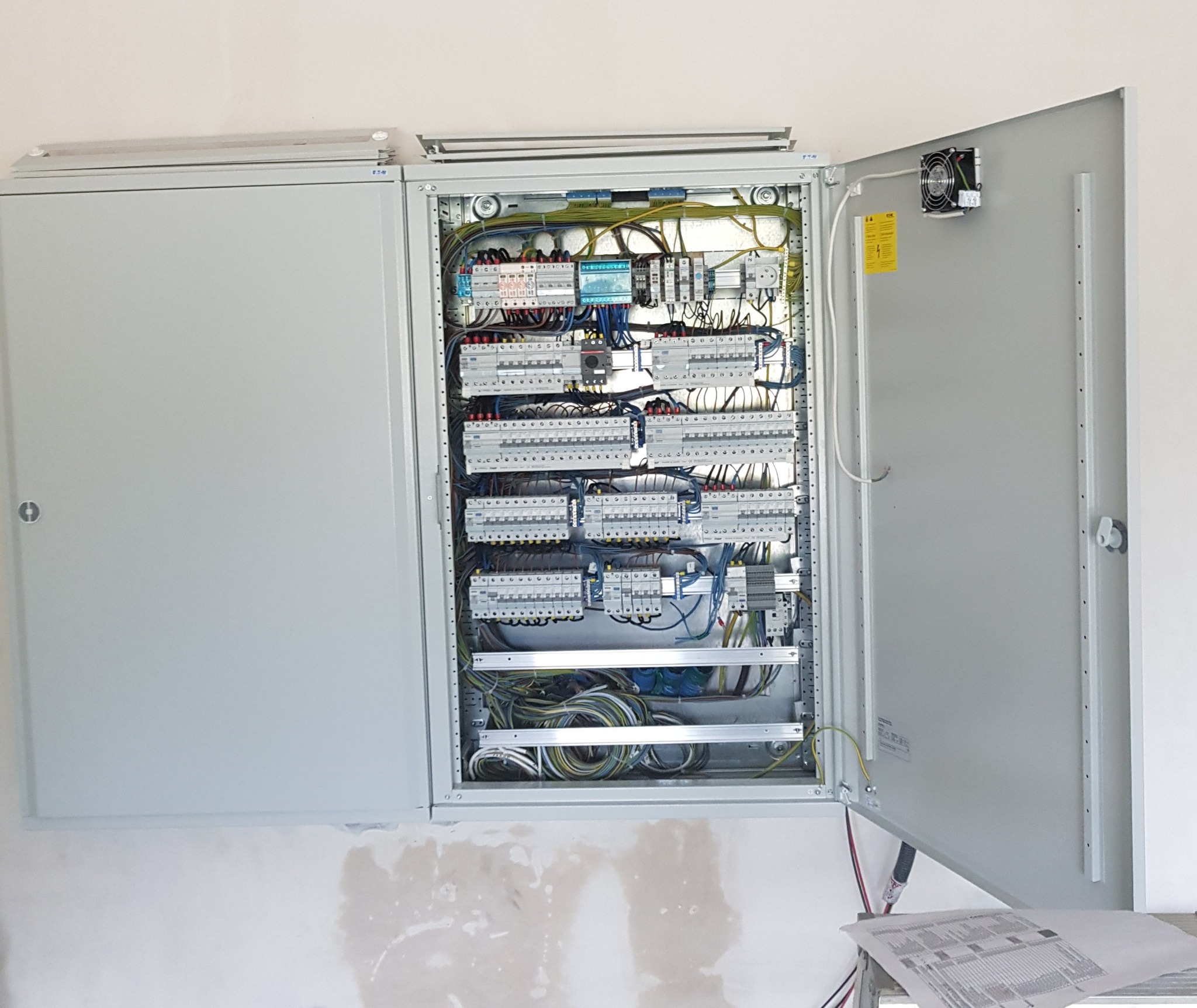 Otwarta skrzynka rozdzielcza z widocznymi zabezpieczeniami, przewodami i elementami montażowymi na białej ścianie. Na drzwiach wentylator i schemat.