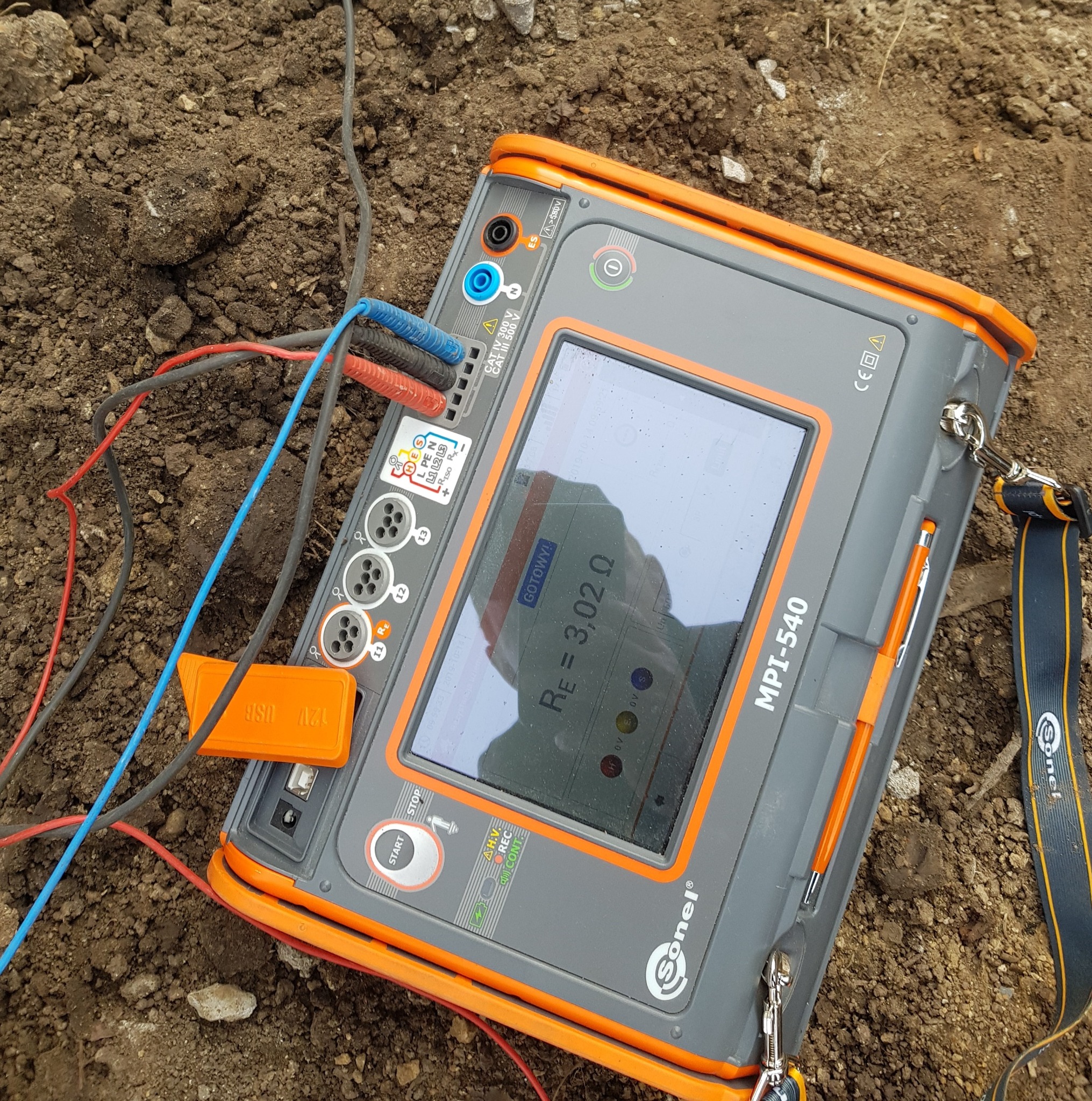 Miernik Sonel MPI-540 na ziemi z podłączonymi przewodami, wskazujący rezystancję 3,02 Ω. Urządzenie używane do pomiarów elektrycznych, widoczne złącza i ekran.