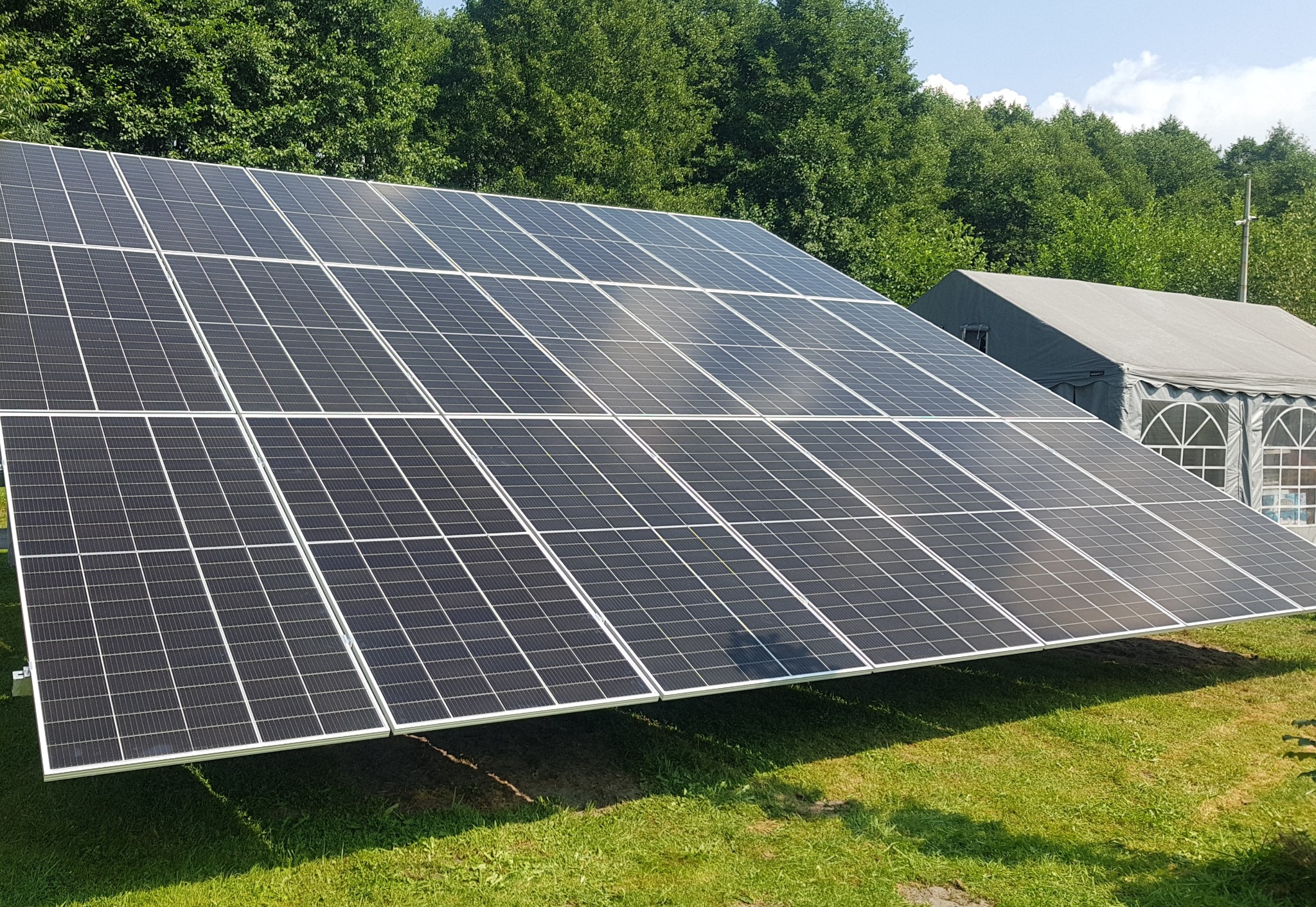 Panel fotowoltaiczny na trawie, z namiotem w tle. Widoczne moduły słoneczne w regularnym układzie, zainstalowane na zielonej powierzchni.