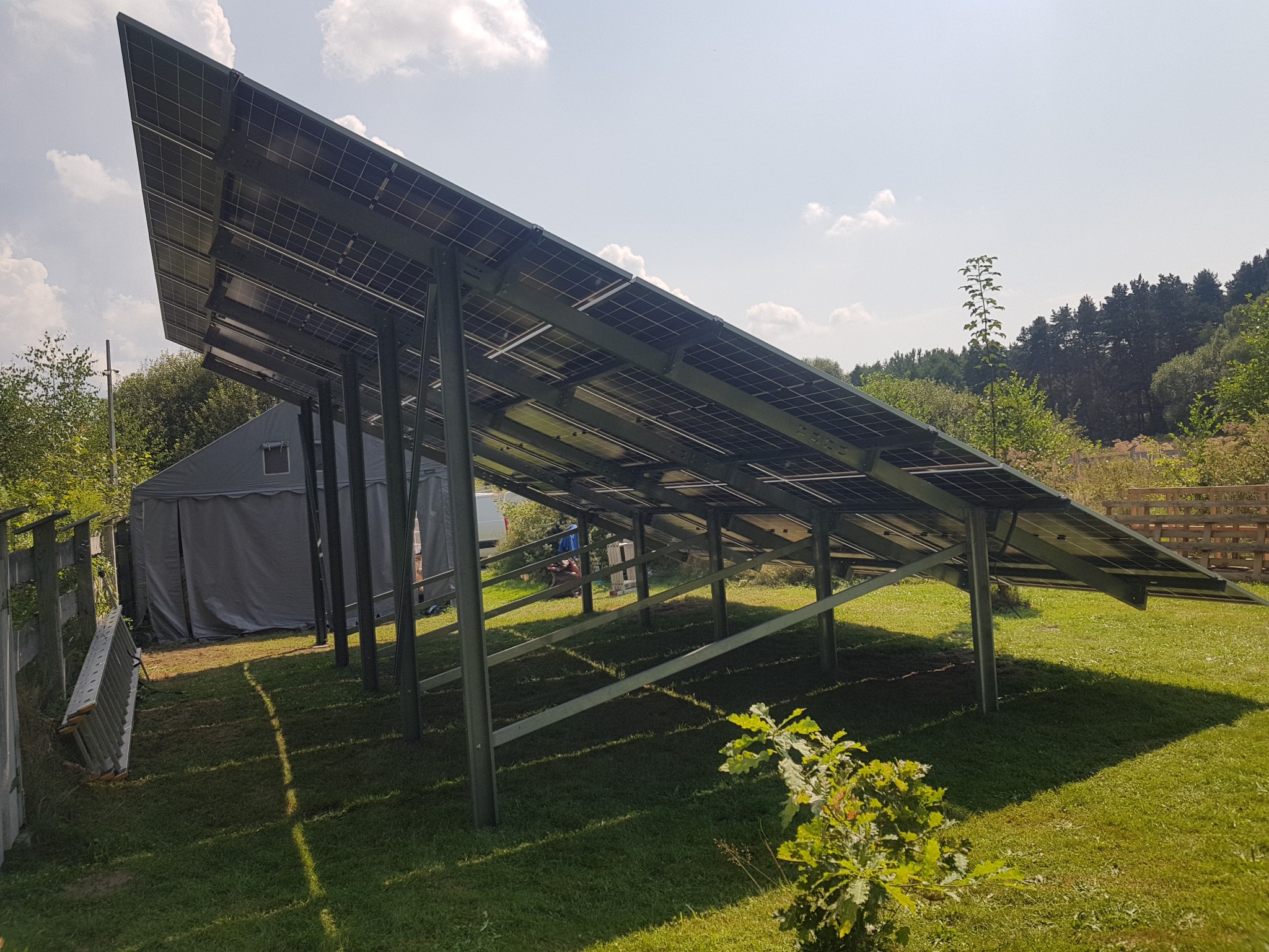 Nisko kątowe ujęcie paneli fotowoltaicznych na metalowej konstrukcji w ogrodzie, z widocznym namiotem i drabiną w tle, w słoneczny dzień.