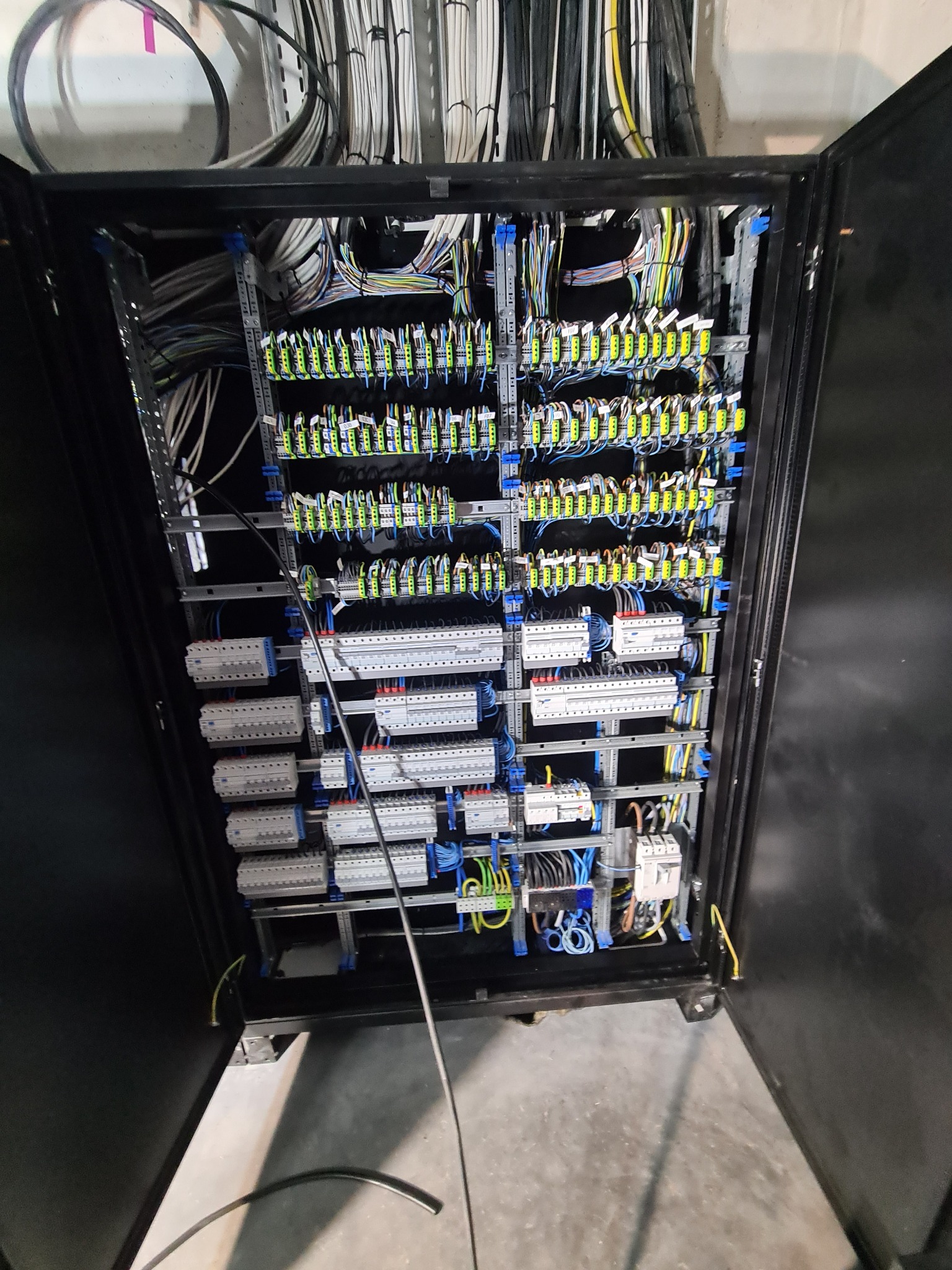 Otwarta szafa sterownicza z licznymi przewodami, listwami zaciskowymi, zabezpieczeniami i szynami montażowymi. Widoczne kable w różnych kolorach ułożone wewnątrz szafy.