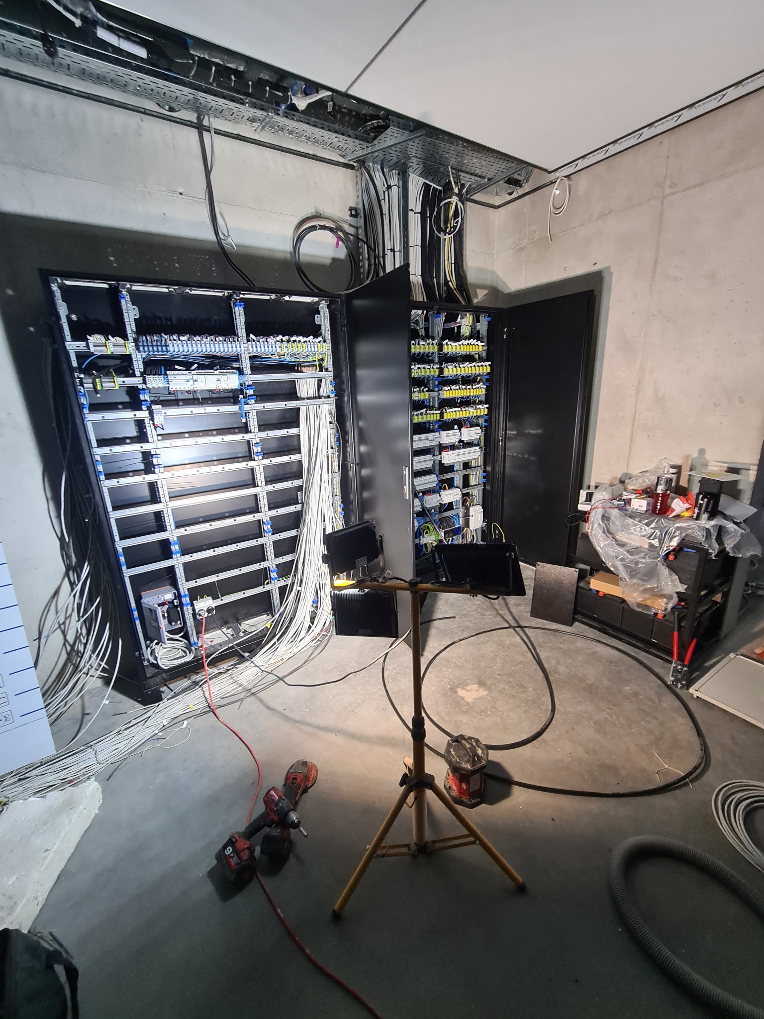 Rozdzielnice elektryczne w trakcie montażu, widoczne liczne przewody, narzędzia i oświetlenie robocze w surowym wnętrzu. Etap prac instalacyjnych.