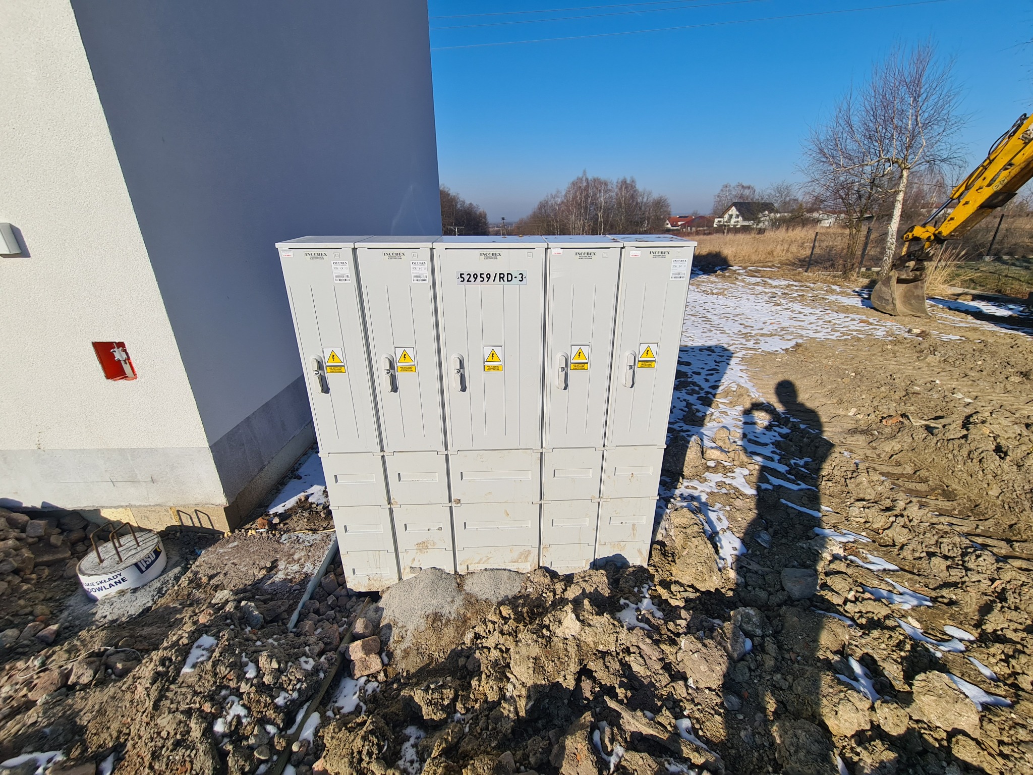 Biała, modułowa stacja transformatorowa Inoccbox 52959/RD-3, z żółtymi znakami ostrzegawczymi, usytuowana na placu budowy z koparką w tle.